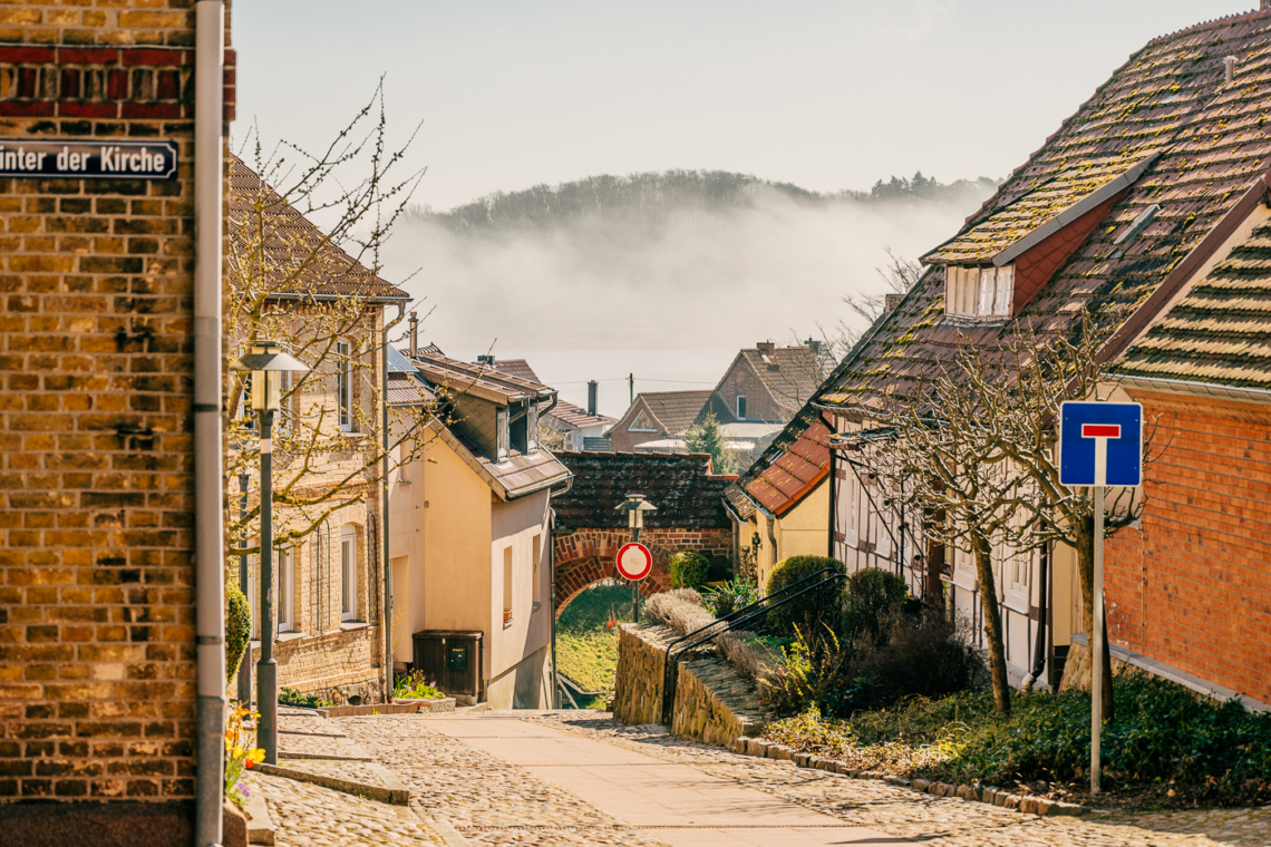 Altstadt Sternberg, Blick durch die Straße Unter der Kirche auf das kleine Stadttor mit Nebel über dem See dahinter.