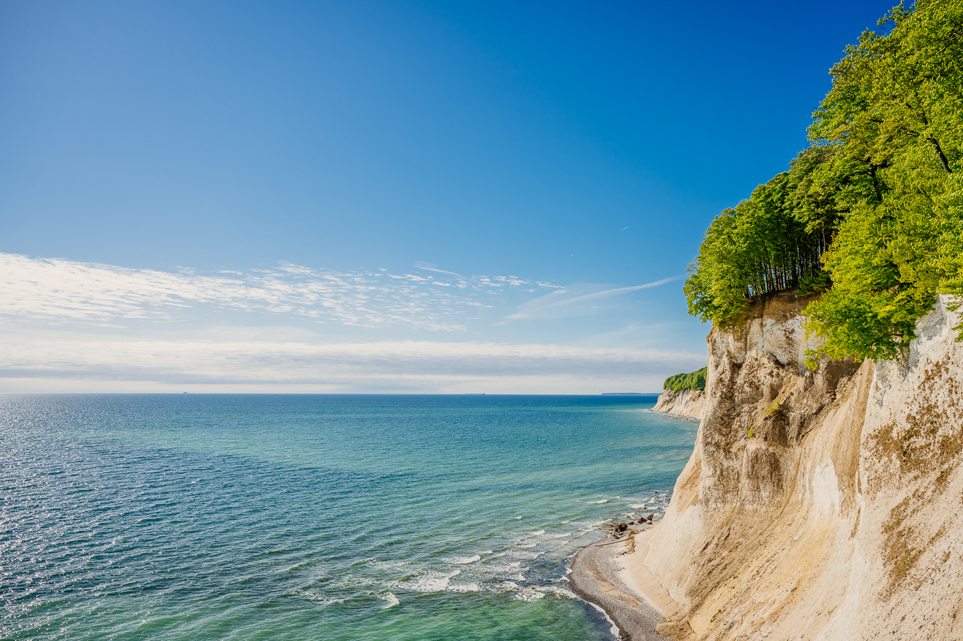 Weite Kreideküste von Jasmund mit bewaldeten Steilklippen, schmalem Strand und ruhiger Ostsee unter hellem Frühlingshimmel.