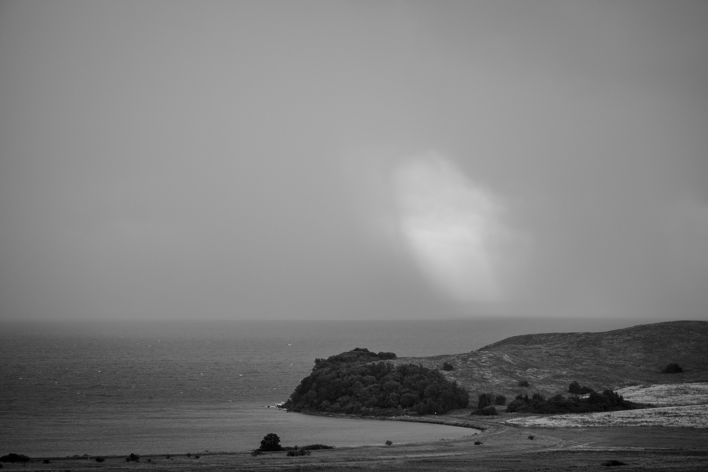 Ein Regenschauer zieht über Meer und Küstenhügel des Mönchguts, während ein heller Wolkenstreifen den Horizont durchbricht.