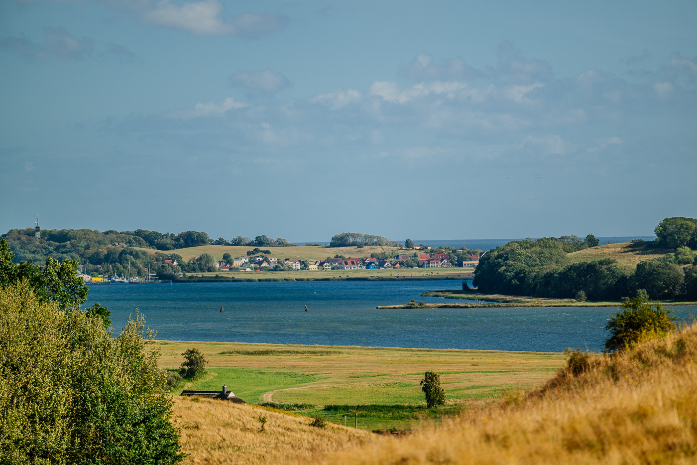Blick von den Zickerschen Bergen über das Wasser nach Thiessow auf Mönchgut, eingerahmt von spätsommerlichen Wiesen und Bäumen.